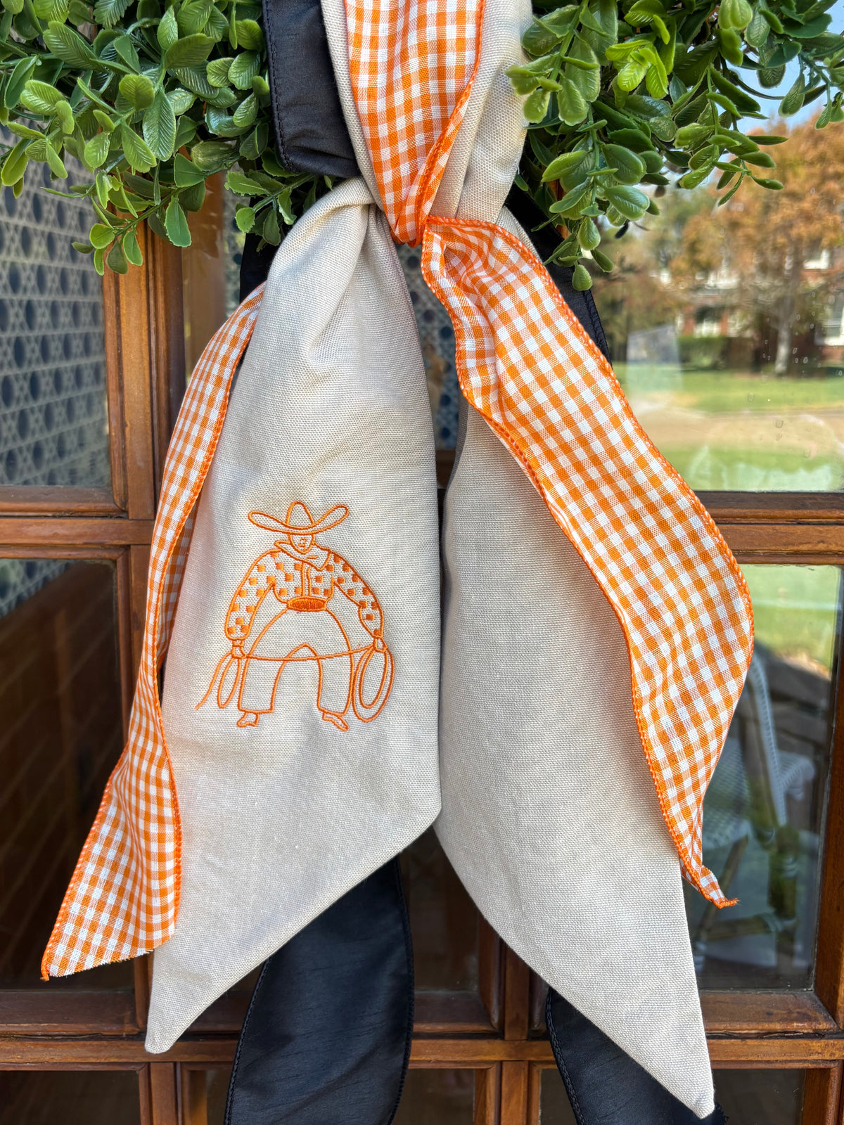 Linen wreath sash with orange embroidered western cowboy, orange gingham ribbon and black ribbon tied around a green wreath